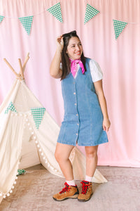 Woman wearing a light wash V-neck button-front denim dress over a white tee, styled with a pink paisley bandana, white socks, and brown lace-up hiking boots, posing in front of a pink backdrop with a white teepee