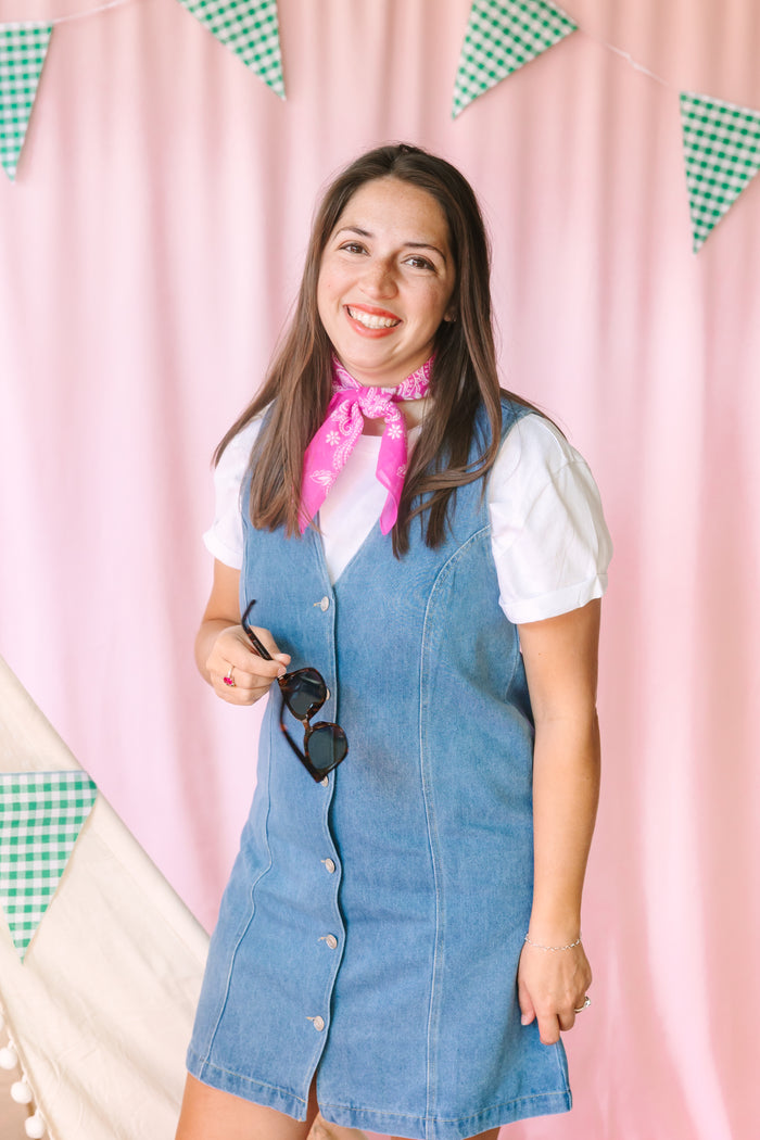 Smiling woman in a sleeveless light wash denim button-front dress layered over a white short-sleeve tee with a pink paisley bandana tied at the neck, holding sunglasses