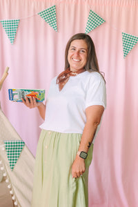 Smiling woman in white t-shirt, green midi skirt, and rust neck scarf holding a children's camping activity book, standing in front of a pink backdrop with green gingham pennant banner decorations