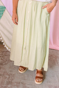 Close-up of a sage green button-front maxi skirt with side pocket and flowy hem, styled with brown platform sandals