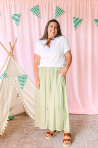 Woman smiling in a white short-sleeve V-neck tee and sage green button-front maxi skirt with side pockets, styled with a rust bandana and brown platform sandals, pink backdrop with green gingham pennant banner