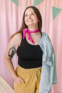 Woman smiling and looking up, wearing a black ribbed tank top, mustard yellow drawstring pants, a pink bandana tied at the neck, and a light wash denim jacket draped over one shoulder, against a pink backdrop