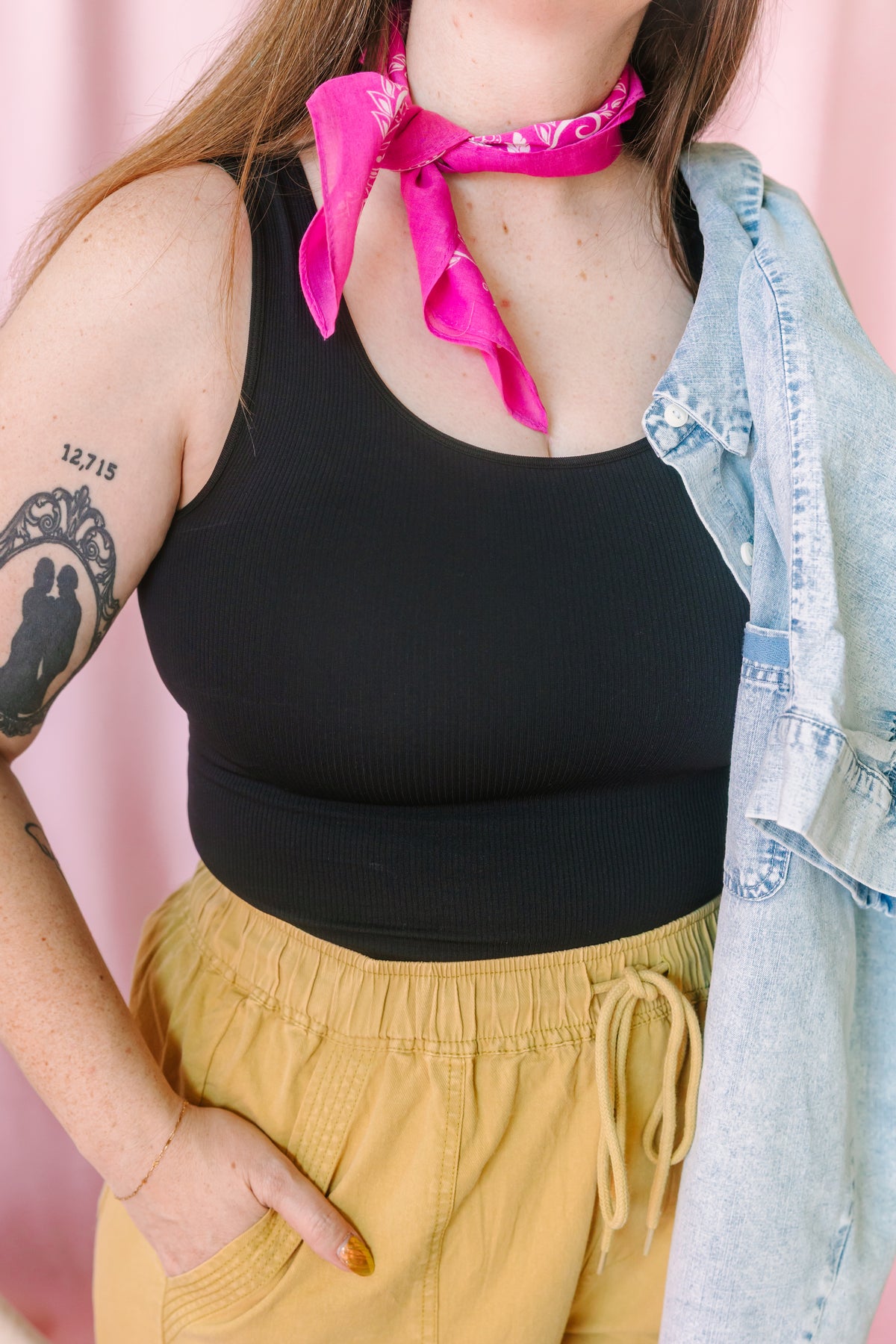 Close-up of a woman wearing a black ribbed tank top, mustard yellow drawstring pants, and a pink paisley bandana tied at the neck, with a light wash denim jacket draped over one shoulder