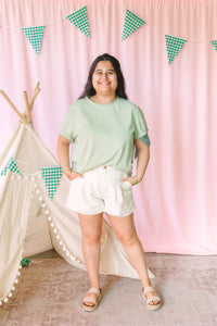 A woman stands in front of a pink curtain backdrop with green gingham bunting and a small fabric teepee. She is wearing a light sage green short-sleeve t-shirt, white shorts, and beige sandals.