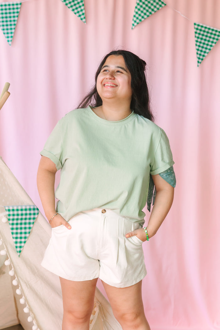 A woman wearing a light sage green short-sleeve t-shirt and white shorts, standing in front of a pink curtain backdrop decorated with green and white gingham bunting.