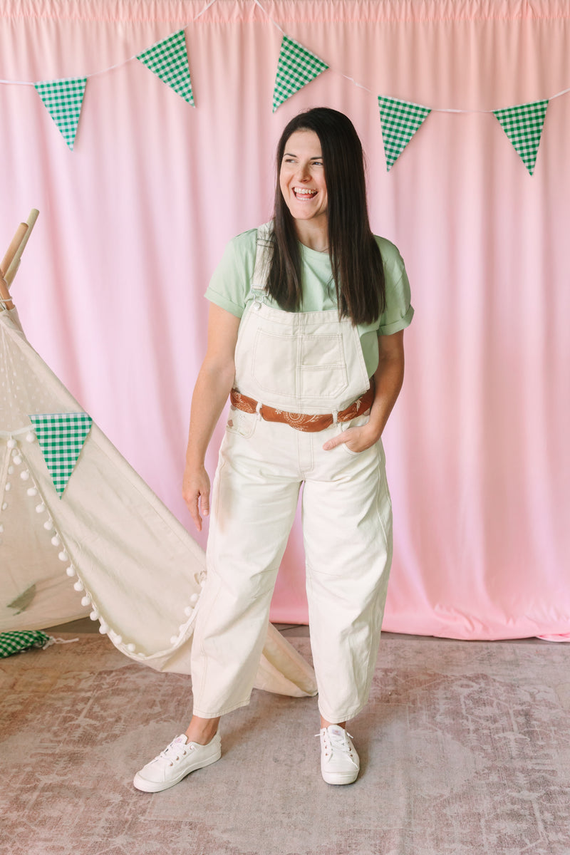 A woman wearing cream-colored overalls over a sage green t-shirt, styled with a brown bandana through the belt loops and white sneakers. 