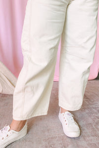 A close-up of a person's legs wearing  cream cropped wide-leg pants and white canvas sneakers, standing against a pink backdrop.