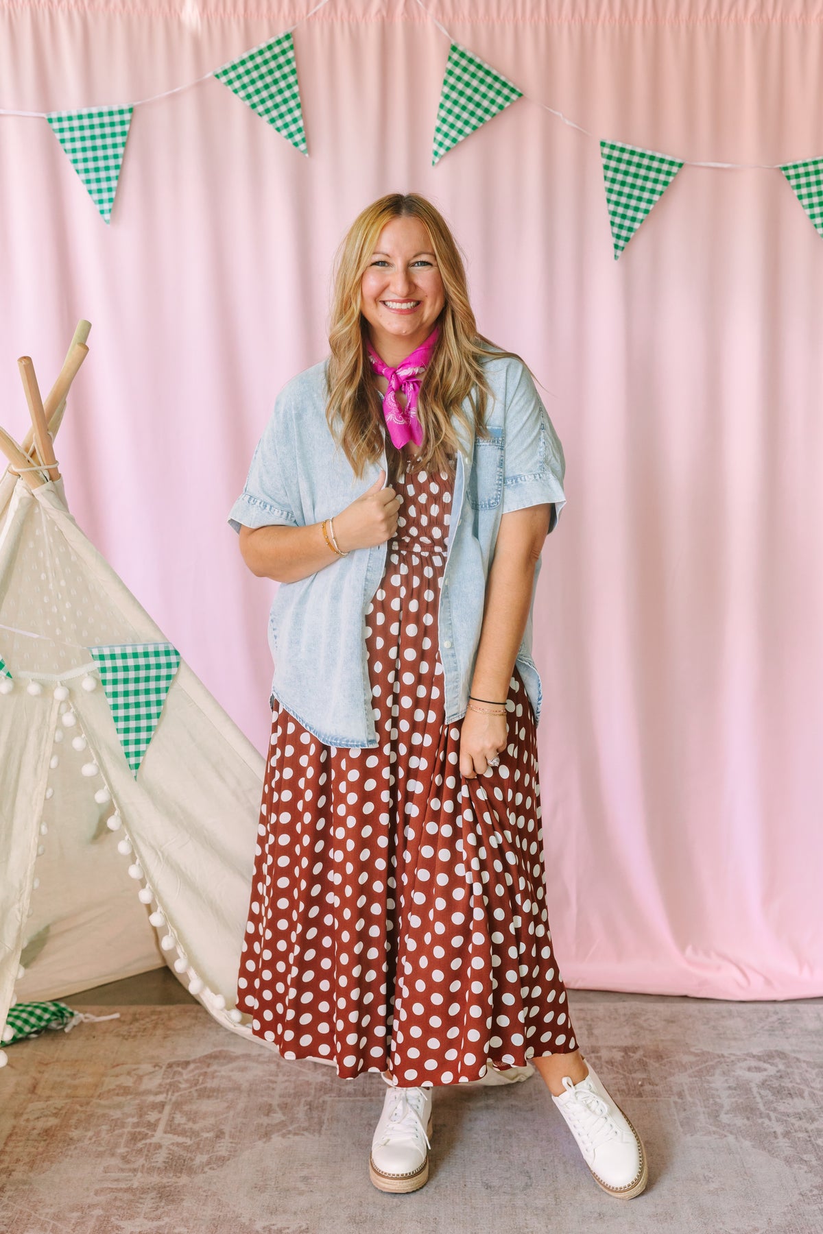 Woman wearing a rust brown polka dot midi dress layered with a light wash short-sleeve denim shirt and pink bandana, styled with white platform sneakers