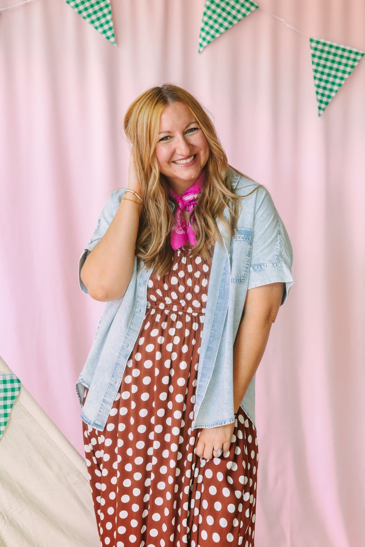 Woman in brown polka dot dress with denim vest and pink bandana scarf, smiling against pink backdrop