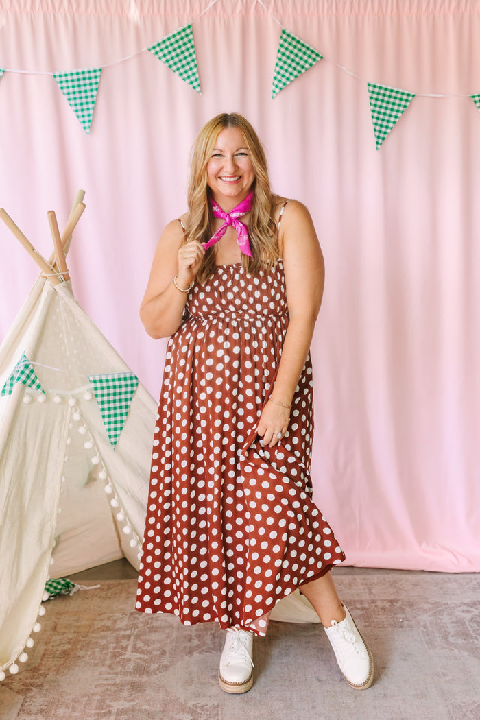 Full-length view of a rust brown and cream polka dot midi dress with smocked bodice and tiered skirt, styled with white platform sneakers and a pink bandana