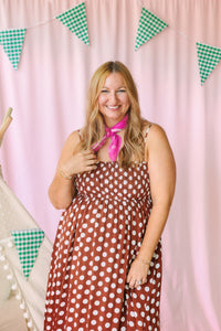 Woman smiling in a rust brown polka dot smocked midi dress with spaghetti straps and a pink bandana scarf, pink backdrop with green gingham pennant banner