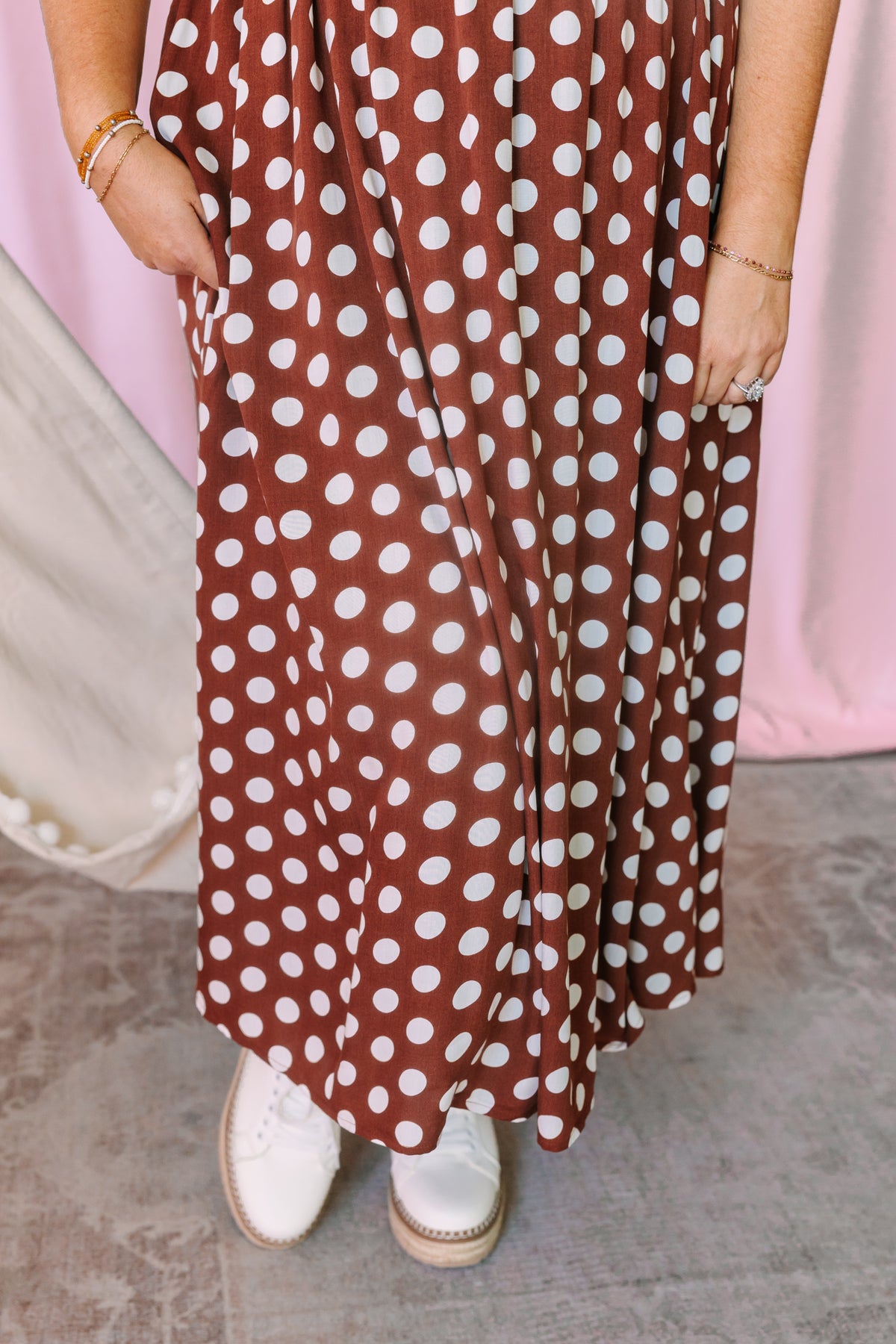 Close-up of the tiered skirt on a rust brown and white polka dot midi dress showing side pockets and flowing hem, styled with white platform sneakers