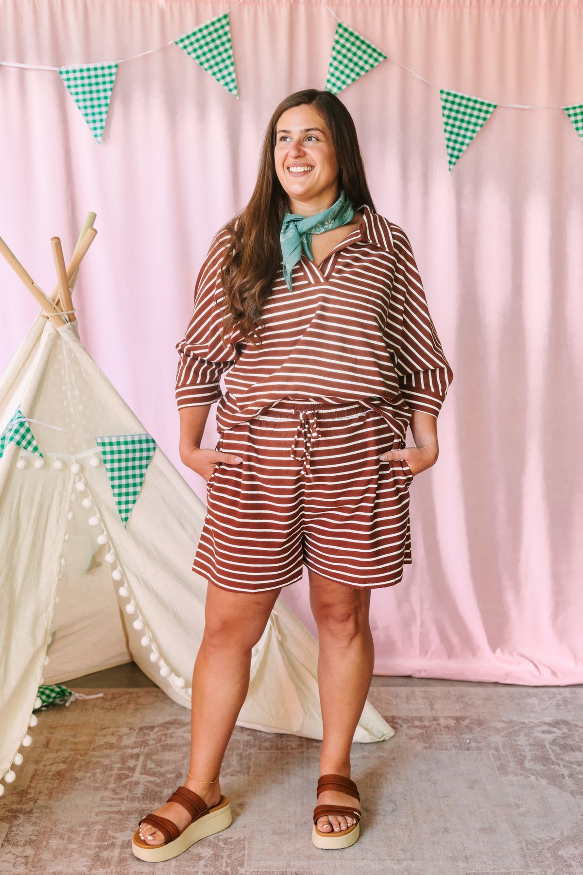Woman smiling in a rust brown and cream striped matching set with V-neck top and drawstring shorts, styled with a teal bandana and brown platform sandals, pink backdrop with green gingham pennant banner