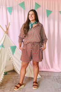 Woman smiling in a rust brown and cream striped matching set with V-neck top and drawstring shorts, styled with a teal bandana and brown platform sandals, pink backdrop with green gingham pennant banner