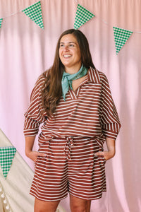 Woman laughing in a rust brown and cream striped matching set with V-neck collared top and drawstring shorts, styled with a teal bandana, pink backdrop with green gingham pennant banner
