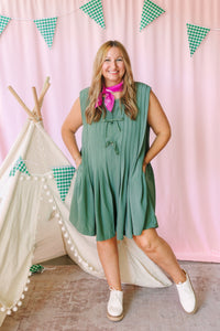 Woman wearing sage green pleated sleeveless romper with tie front and pink bandana scarf, styled with white sneakers in a playful pink backdrop