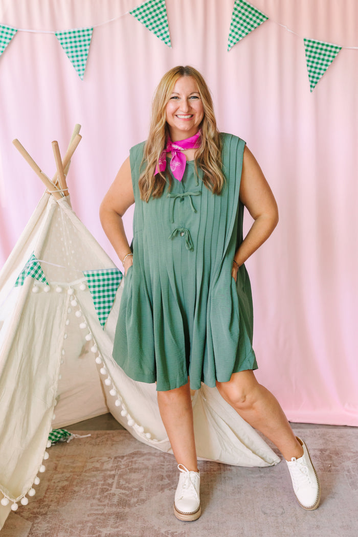 Woman wearing sage green pleated sleeveless romper with tie front and pink bandana scarf, styled with white sneakers in a playful pink backdrop