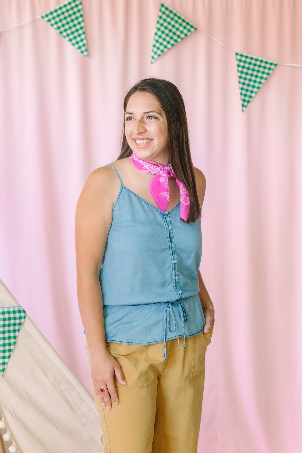 Woman smiling in a light blue chambray button-front cami top with drawstring hem, pink paisley bandana tied at neck, and khaki wide-leg pants, styled in front of a pink backdrop with green gingham pennant banner