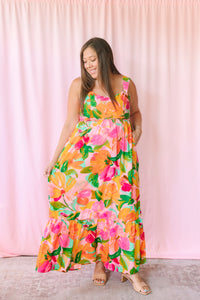 Woman wearing a vibrant tropical floral maxi dress with orange, pink, and green blooms, tiered ruffle hem, and square neckline
