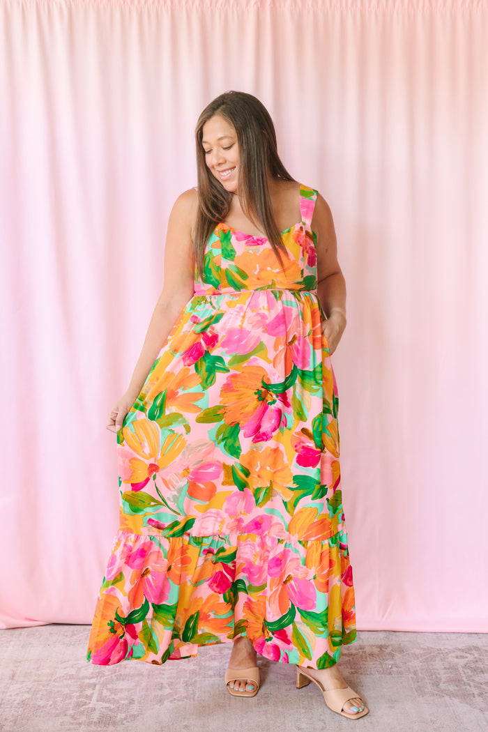 Woman wearing a vibrant tropical floral maxi dress with orange, pink, and green blooms, tiered ruffle hem, and square neckline