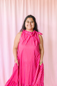 Woman smiling in a hot pink and red striped sleeveless maxi dress with ruffle neckline and bow tie detail, hands in pockets