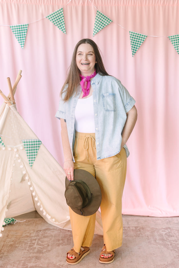 Woman wearing a white seamless ribbed tank top layered under a light denim shirt with a pink bandana, yellow wide-leg pants, and platform sandals at a camp-themed photo shoot