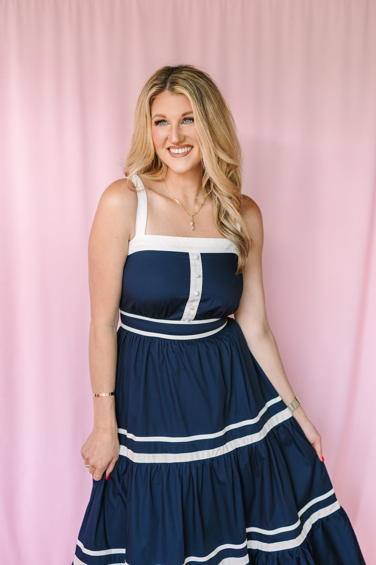 Smiling woman in navy tiered maxi dress with white contrast trim and button detail, pink backdrop