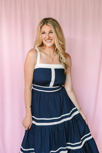 Smiling woman in navy tiered maxi dress with white contrast trim and button detail, pink backdrop