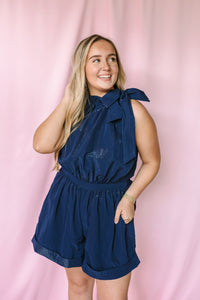 Navy sleeveless romper with bow tie halter neckline, elasticized waist, and shorts — front view on model