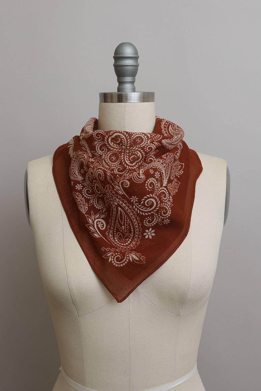 Rust brown paisley print bandana tied around a dress form mannequin, featuring intricate white floral and teardrop paisley pattern
